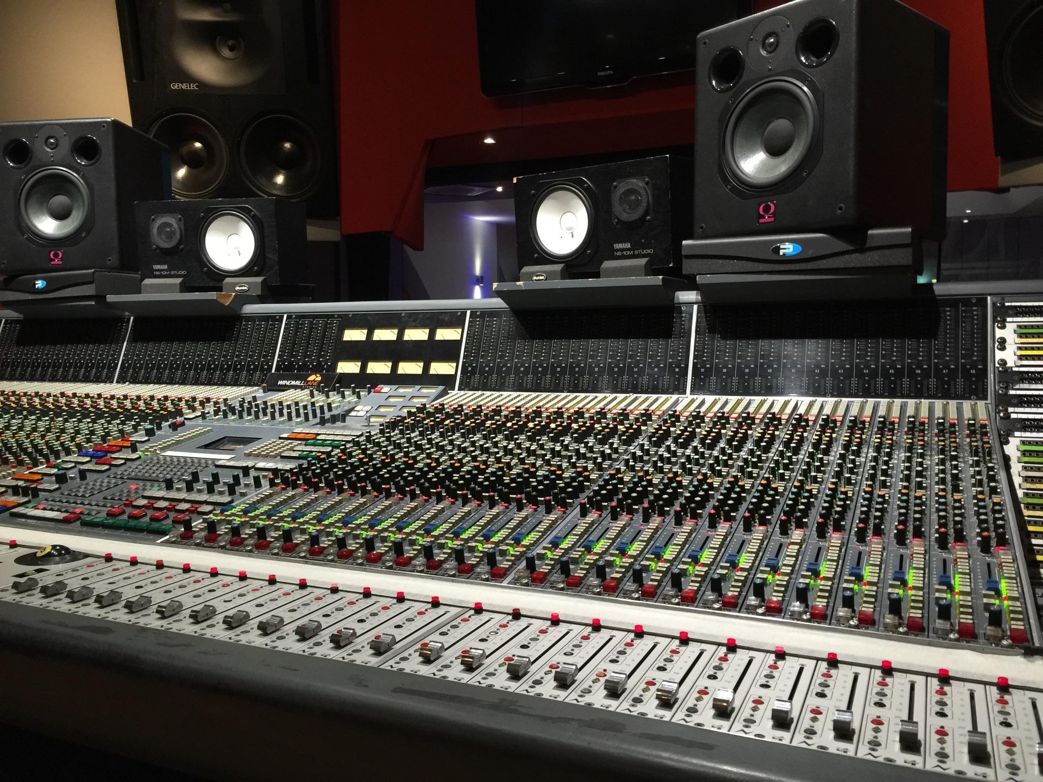 A large analog mixing console under a row of studio monitors in a professional control room