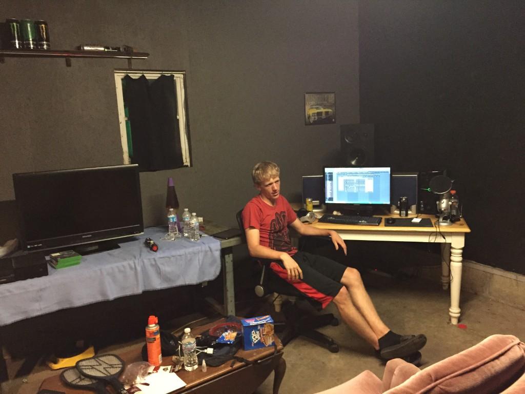 A small room with black walls, a desk with a monitor, speakers, and a microphone, and a friend leaning back in a desk chair