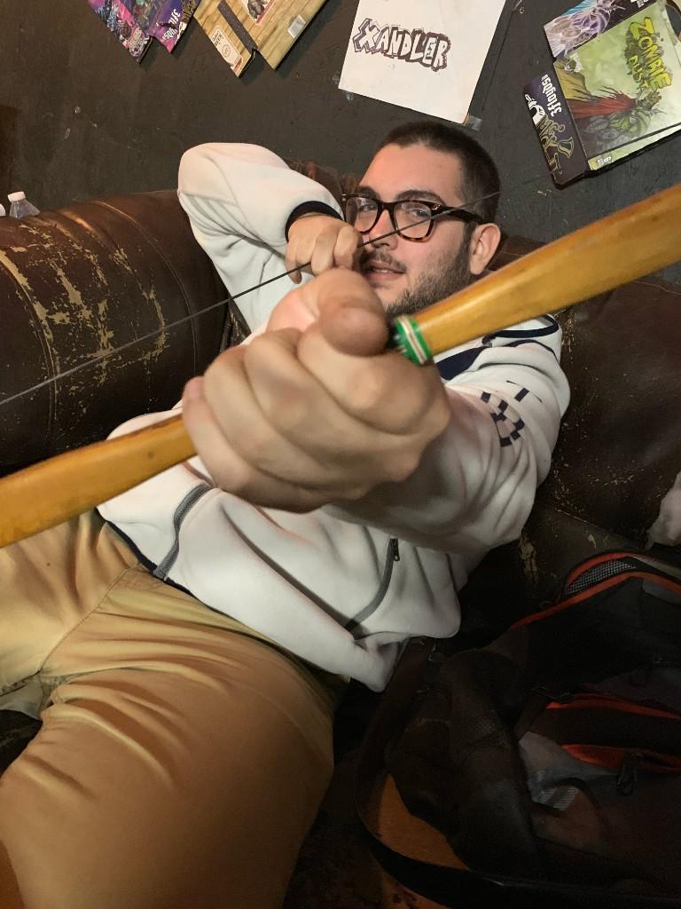A friend on the couch aiming a wooden bow toward the camera