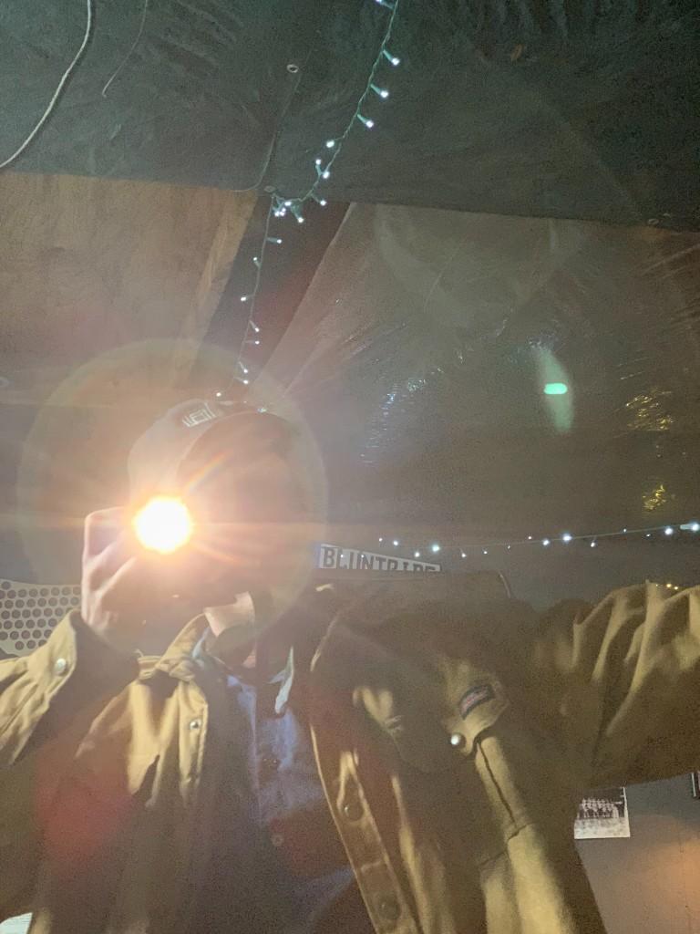 A mirror selfie with the phone flash flaring, taken in a tan work jacket and cap under the string lights in the garage