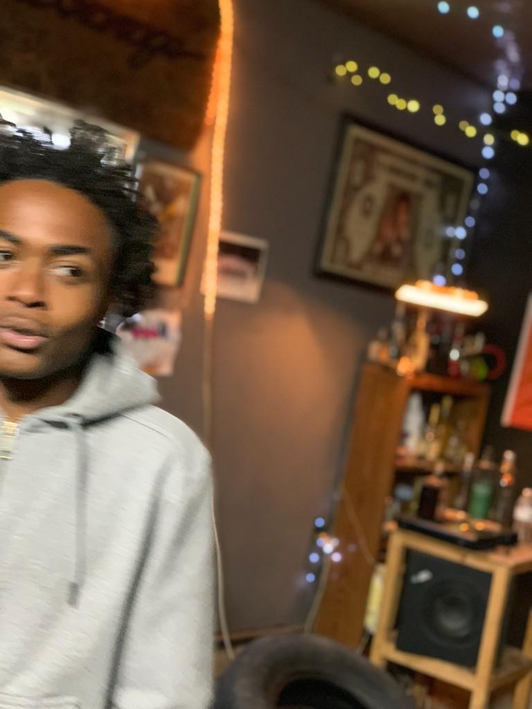 An artist in a gray hoodie in the garage, with string lights and a shelf of bottles behind him
